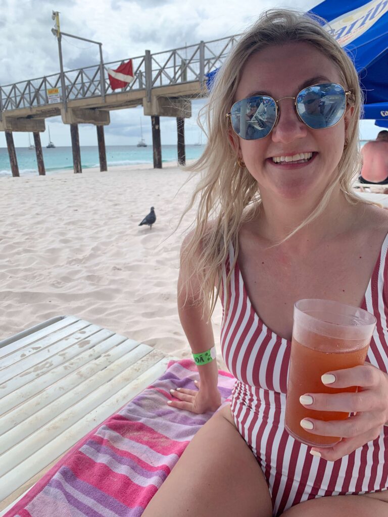 Woman sat on a sunlounger holding a rum punch, wearing sunglasses on the beach with a wooden pier in the background