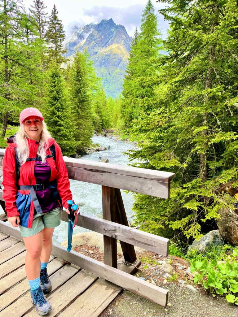 Woman wearing outdoor clothes stood on a wooden bridge over a river with fir trees either side