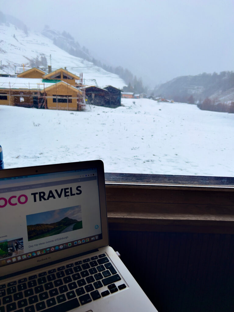 Laptop open to CocoTravels.co.uk with a view of a snowy landscape out of the window.