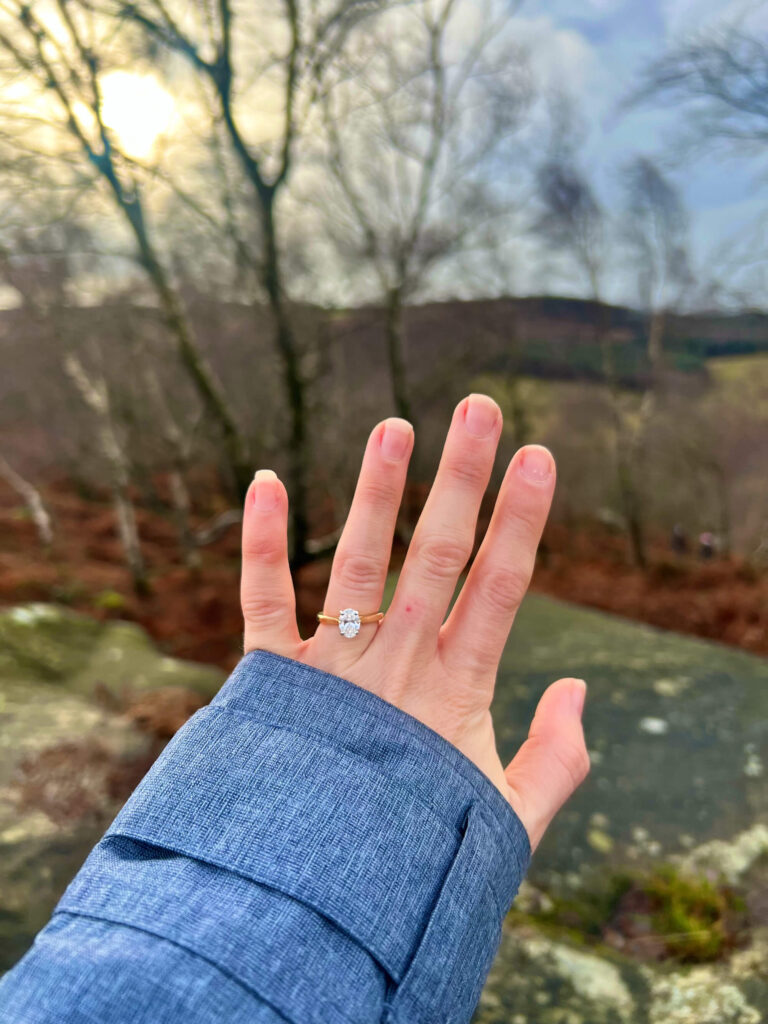 A woman holds her left hand wearing her engagement ring out over a view from Birchen Edge.