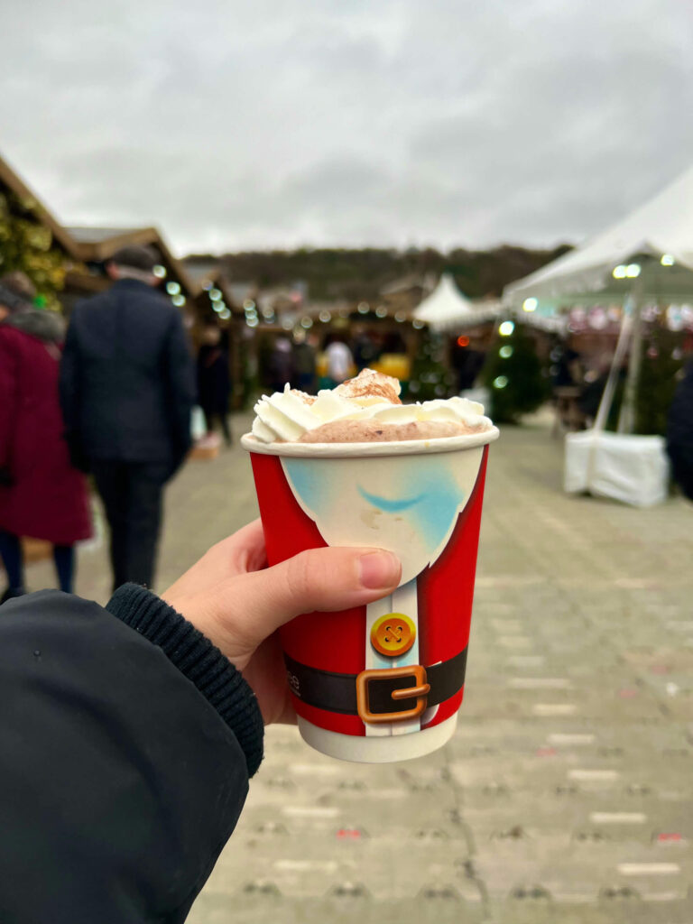 A hand holding a Christmas cup in front of stalls at Chatsworth Christmas market.