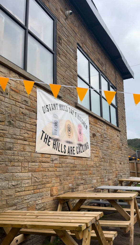 The Distant Hills Brewing sign with orange bunting and wooden picnic tables in front of it.