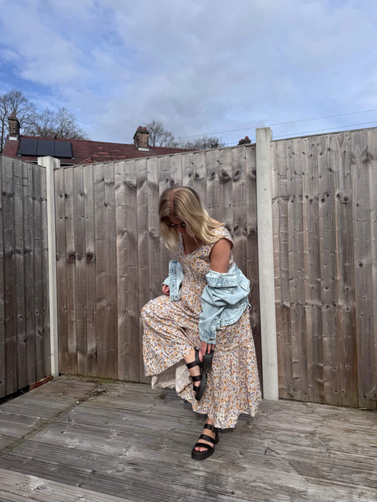 Sam wearing a floral midi dress and faded denim jacket, fixing her shoe.
