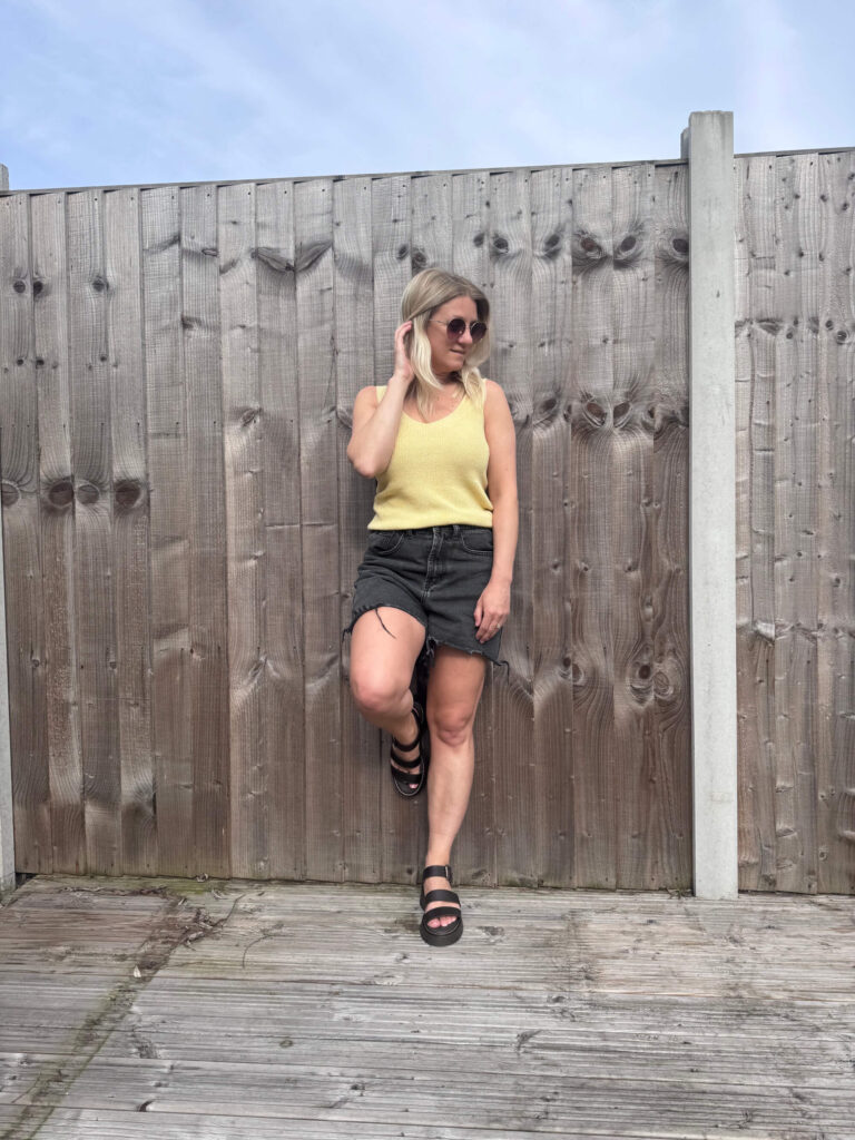 Sam wearing a yellow top and black shorts leaning against a wooden fence.